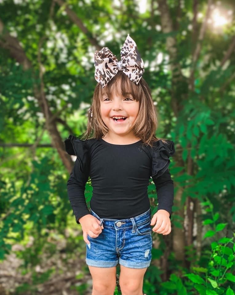 toddler in woods outside in black ruffle leotard jean shorts and large cow bow